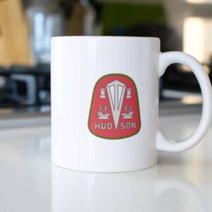 Classic white 11oz ceramic mug emblazoned with the red and green Hudson Motor Car logo, displayed on a kitchen countertop.