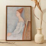 Painting by Amedeo Modigliani of Jeanne Hébuterne, depicted with a characteristic elongated neck and a contemplative gaze. Her figure is adorned in a softly draped garment, with auburn hair styled in an updo, set against a muted backdrop that emphasizes the subject's ethereal beauty and Modigliani's distinctive use of form and color.