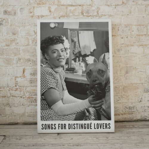 A framed poster on a wall shows a smiling Billie Holiday with a flower in her hair holding a dog, titled Songs for Distingue Lovers. Objects on a shelf include a camera, plant, books, laptop, and glass vase.