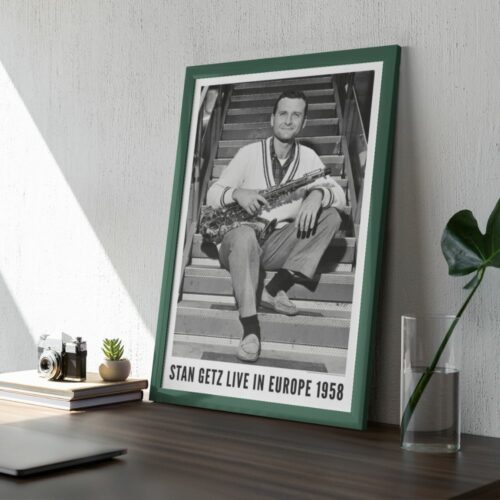 A framed poster on a wall depicts a smiling man seated on steps, holding a saxophone, with text Stan Getz Live in Europe 1958. Beside the poster, a camera, a plant, books, a laptop, and a glass vase sit on a shelf.