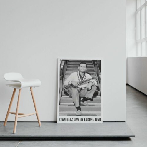 A framed poster on a wall depicts a smiling man seated on steps, holding a saxophone, with text Stan Getz Live in Europe 1958. Beside the poster, a camera, a plant, books, a laptop, and a glass vase sit on a shelf.