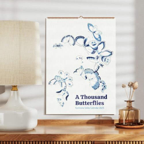 Wall calendar cover with blue and white watercolor butterflies design titled 'A Thousand Butterflies Kamisaka Sekka Calendar 2025' in a home setting with a lamp and vase.