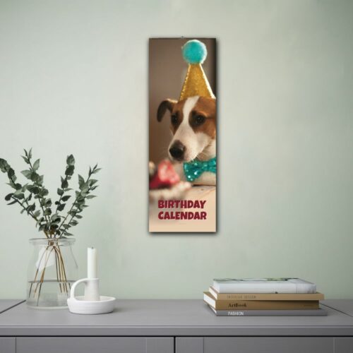 Wall calendar featuring a dog wearing a gold party hat and blue bow tie. The calendar is labeled Birthday Calendar and is displayed on a wall beside a lamp and an open book, creating a cozy setting.
