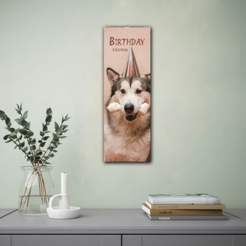 Wall calendar featuring a husky dog wearing a party hat and holding a bone-shaped chew toy in its mouth. The calendar is labeled Birthday Calendar and is displayed on a wall beside a lamp and an open book.