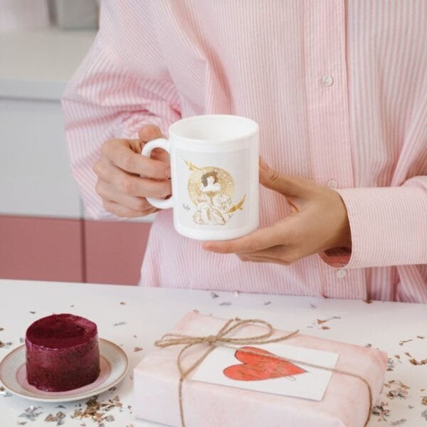Person holding mug with cake and gift nearby.