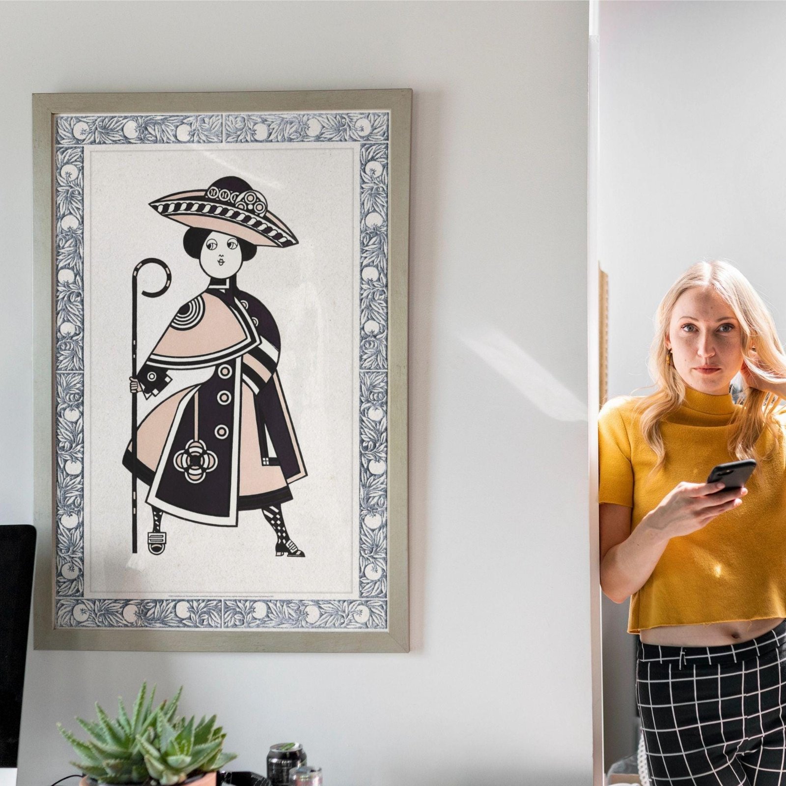 Woman holding phone beside framed art on wall.